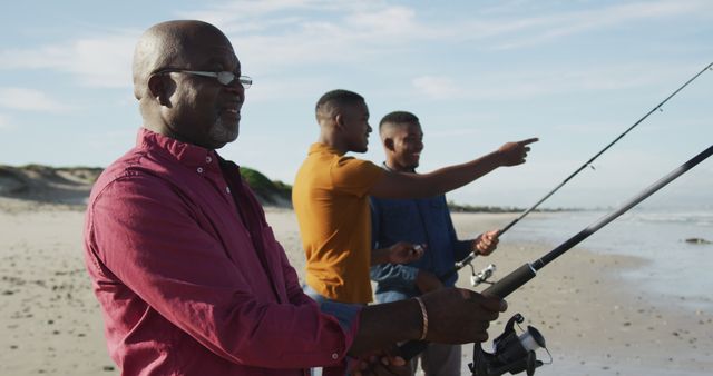 African american senior father and twin teenage sons standing on a beach fishing and talking. healthy outdoor family leisure time together.