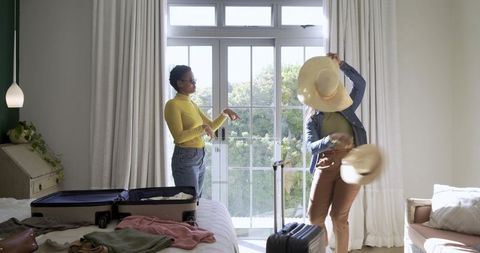 Female friends trying hats in sunlit bedroom with open suitcase