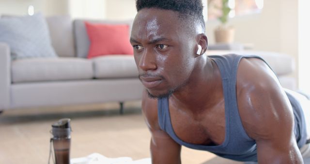 Image depicts an African American man engaged in a workout at home, showcasing his determination and focus. He is wearing earbuds and appears to be sweating, indicating a vigorous exercise session. A water bottle is visible nearby, emphasizing the importance of hydration. Suitable for use in fitness-related content, home workout guides, healthy lifestyle promotions, and mental focus themes.