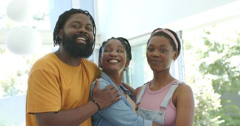Smiling family hugging in sunlit modern living room, casual parents and teen daughter