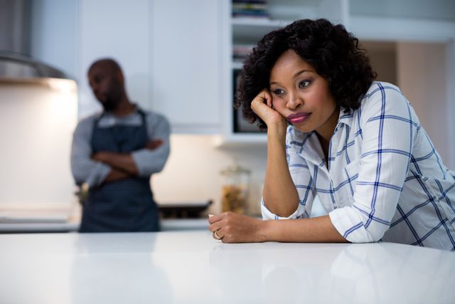 African American couple experiencing relationship problems in modern kitchen. Woman looks sad and thoughtful while man stands in background with arms crossed. Useful for topics on relationships, conflict resolution, emotional stress, and family dynamics.