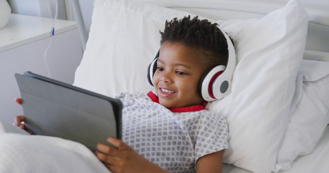 Happy african american boy patient using tablet and wearing headphones in bed at hospital. Medicine, healthcare, lifestyle and hospital concept.