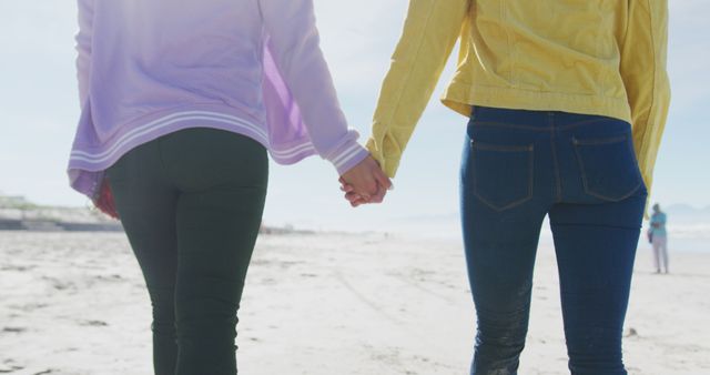 Midsection of diverse female gay couple holding hands walking along beach. female friends bonding at the beach.