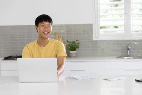 Asian young man working on laptop in modern kitchen smiling while checking papers