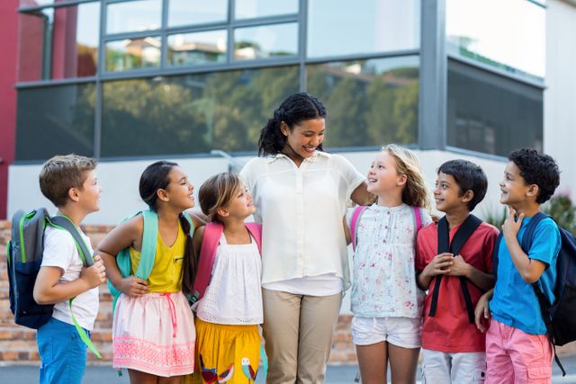 Female teacher standing with a group of diverse children outside a school building. They are all smiling and wearing backpacks, indicating a school setting. This image can be used for educational materials, school advertisements, or articles about teaching, childhood education, and student-teacher relationships.