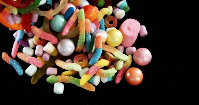 Collection of diverse colorful sweets including gummy worms, marshmallows, and sugar-coated candies on a stark black background. Ideal for marketing materials related to candies, dessert recipes, or children's parties. This vibrant image captures attention and can be great for advertisements, social media posts, and editorial content promoting fun and sweetness.