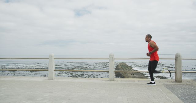 Fit young man jogging along a seaside promenade with ocean in background. This image can be used for promoting fitness, outdoor activities, healthy lifestyle, urban exercise environments, and athletic wear. Excellent for use in advertisements for sports apparel, fitness programs, and wellness blogs.