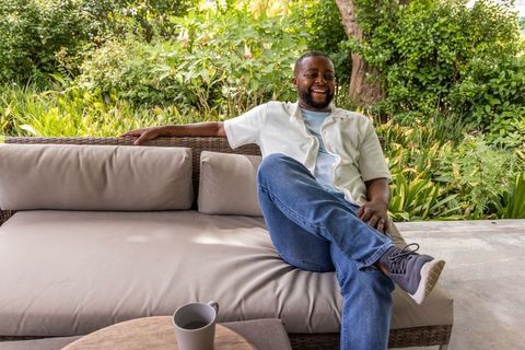 African man relaxing on outdoor wicker sofa at lush garden patio, smiling and unwinding