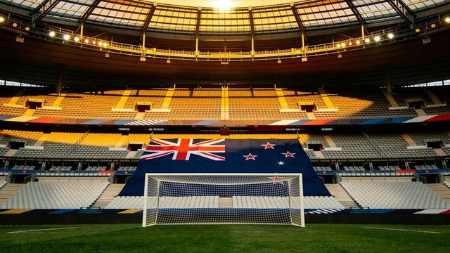 Sunlit stadium interior featuring centered soccer goal with large draped new zealand flag