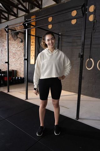 Young woman standing in gym wearing cropped hoodie and cycling shorts, smiling confidently