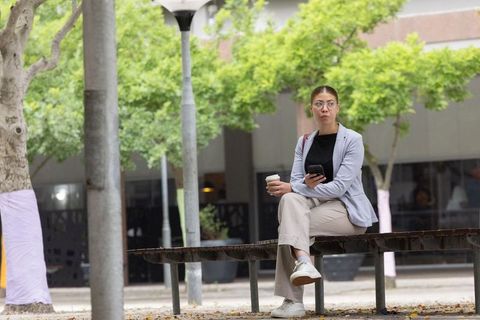 African woman sitting on metal bench holding coffee and checking smartphone in city plaza