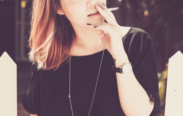 Young woman smoking a cigarette outdoors during daytime. Wearing casual black clothing, long necklace, and wristwatch. Can be used for themes related to smoking, fashion, lifestyle choices, casual scenes, or youth culture.