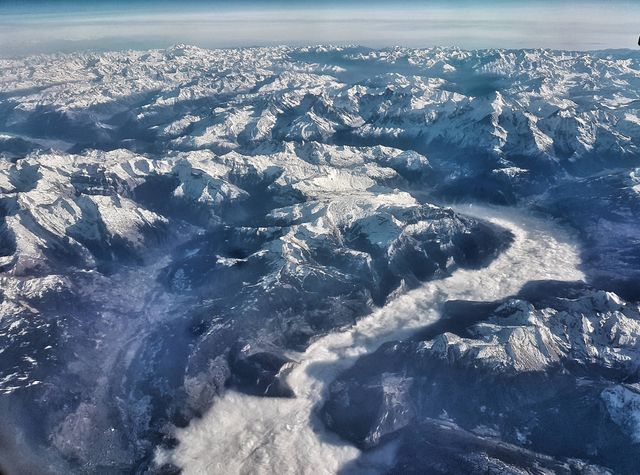 Aerial perspective shows expansive snow-capped mountain range with a winding river valley below, epitomizing nature's winter splendor. Ideal for travel and tourism promotions, adventure sports ads, winter season visitor guides, scenic landscapes, nature lovers' websites, and desktop wallpapers.