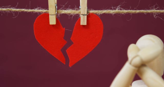 A broken red heart hangs from a clothesline, symbolizing heartbreak or the end of a relationship, with copy space. A wooden figure sits beside it, adding a sense of loneliness or contemplation to the image.