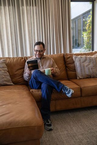 Mature asian man reading on leather sectional holding mug and paperback cozy living room