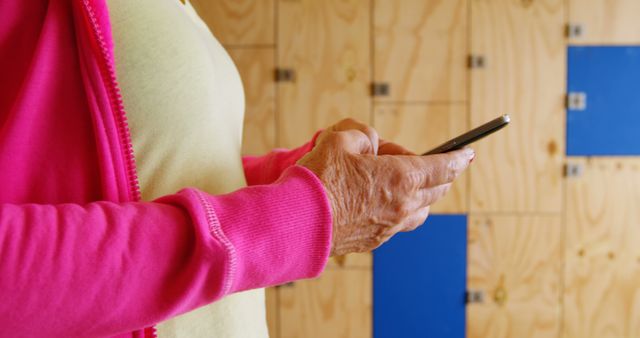Senior woman wearing pink jacket using smartphone close-up in modern environment with wooden background. Suitable for topics on technology adoption among older adults, senior lifestyle, and tech-savvy seniors. Can be used in blogs, articles about elder technology engagement, and promotional materials for senior-friendly technology.