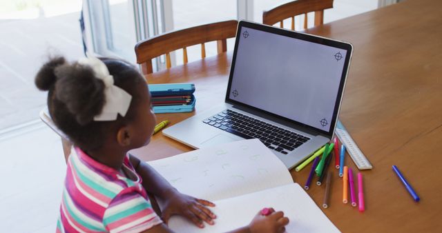This image showcases an African American girl engaged in online learning using her laptop while doing homework. Suitable for articles, blogs, and websites focusing on children's education, homeschooling, online learning tools, and the importance of technology in education. Ideal for promoting virtual education platforms, school supplies, or educational apps.