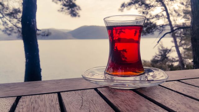 Traditional Turkish tea served in a glass on a wooden table overlooking a scenic lake view with trees. Ideal for promoting travel destinations, wellness blogs, relaxation techniques, or healthy lifestyle campaigns. Suitable for use in articles about Turkish culture, nature retreats, or outdoor activities.