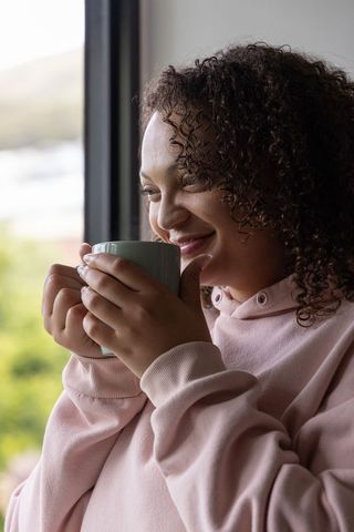 African american woman holding mug by window smiling in pink hoodie cozy morning calm