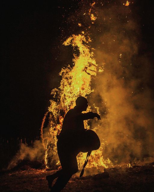 A person seen in silhouette dancing near a large bonfire at night. The fire illuminates the scene with vibrant flames and embers. Use for themes involving celebration, nighttime activities, festive events, or cultural traditions involving fire.