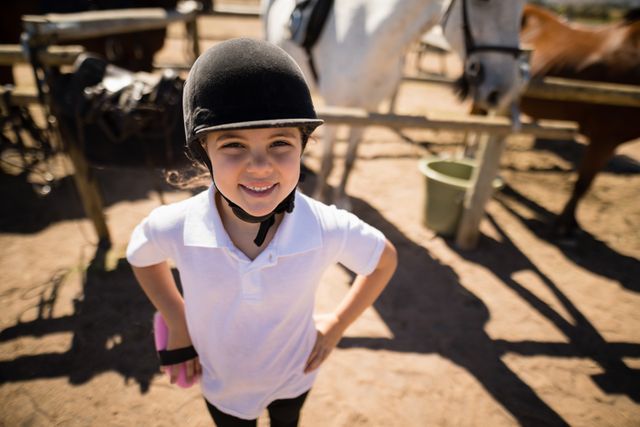 Young girl wearing an equestrian helmet and white shirt, standing with hands on hips at a ranch. Horses and stable equipment visible in the background. Ideal for use in advertisements for equestrian activities, children's outdoor programs, or rural lifestyle promotions.
