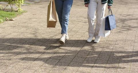 Two women walking with shopping bags on sunny sidewalk casting tree shadows