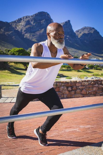 Senior African American man performing stretching exercises outdoors on a sunny day with mountains in the background. Ideal for promoting active senior lifestyles, fitness routines, health and wellness programs, and retirement activities.