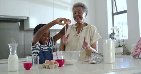 Grandmother guiding granddaughter baking and cracking eggs on bright modern kitchen island