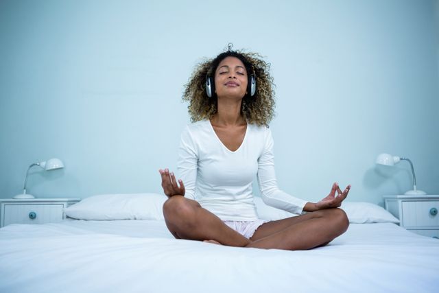 Woman sitting cross-legged on bed, listening to music through headphones while meditating. Ideal for promoting relaxation, mindfulness, self-care, and wellness. Suitable for articles, blogs, and advertisements focused on mental health, tranquility, and indoor lifestyle.
