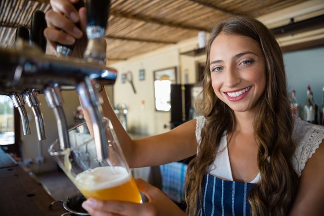 Young woman bartender smiles while pouring a glass of beer from a tap at a restaurant or pub. Suitable for hospitality industry promotions, advertisements for drinking establishments, and features on restaurant services and staff.