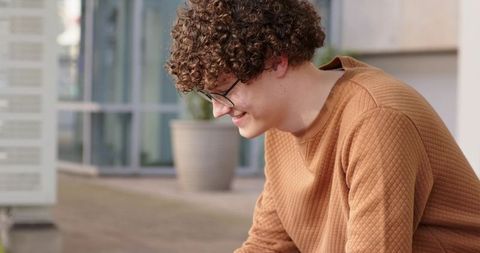 Young man in camel quilted sweater leaning at modern building entrance smiling outdoors