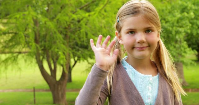 Blonde girl with pigtails stands in green park waving her hand. She smiles warmly, wearing casual clothes, giving a friendly and welcoming vibe. This image is great for use in advertising activities for children's events, friendly community projects, outdoor fun days, or any context involving welcoming gestures or friendly children's themes.
