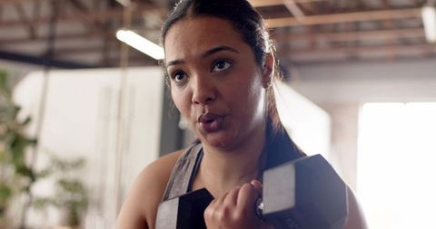 Determined woman lifting dumbbell close to chest in bright gym studio showing strength