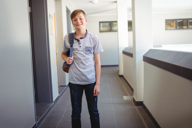Portrait of smiling schoolboy standing with schoolbag in corridor at school