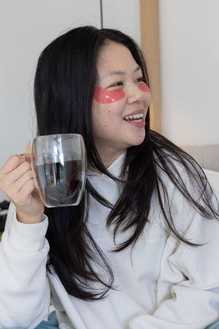 Asian woman enjoying coffee with red under-eye hydrogel patches in cozy minimalist home