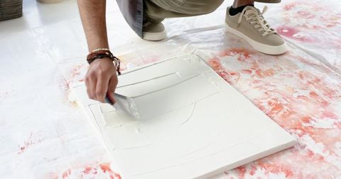 Indian man applying gesso to canvas with palette knife on paint-splattered drop cloth