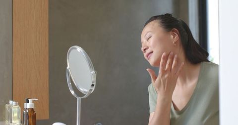 Asian woman applying skincare at vanity with round mirror, pump bottle and soft light