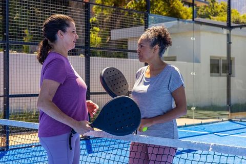 Two diverse women chatting on blue padel court holding paddles and ball