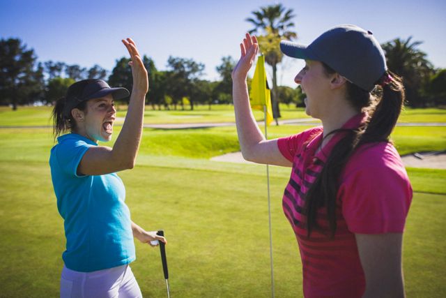 Two women are celebrating a successful moment on a golf course by giving each other a high five. They are dressed in sporty attire and appear to be enjoying a sunny day outdoors. This image can be used to promote sports, active lifestyles, teamwork, and friendship. It is ideal for advertisements, sports magazines, fitness blogs, and social media posts related to golf and outdoor activities.