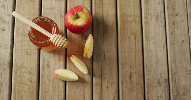 Fresh red apple and jar of golden honey placed on rustic wooden table. A few apple slices scattered. Perfect for use in healthy eating, farm-to-table, autumn themes, Rosh Hashanah, and natural food settings. Favored for illustrating fresh produce and simple snacking choices.