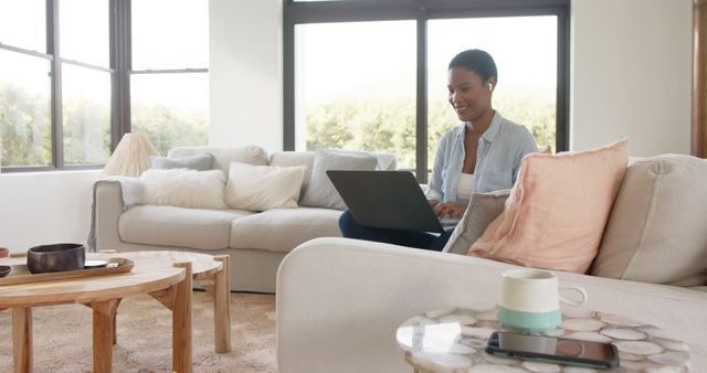 Woman working on a laptop while sitting in a cozy living room. Ideal for themes such as remote work, home office, freelancing, modern lifestyles, and technology. Can be used in articles or advertisements about remote work, home comfort, and balancing professional life with personal life.