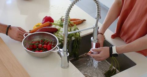 Couple rinsing carrots at modern sink with colander of cherry tomatoes and bell peppers