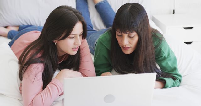 This image shows two young women lying on a bed, engaging with a laptop together in a relaxed manner. One wears a pink sweater while the other is in a green sweater, indicating a casual and comfortable setting. This image can be used for themes related to friendship, study sessions, remote work, online shopping, social media interaction, or leisure activities.