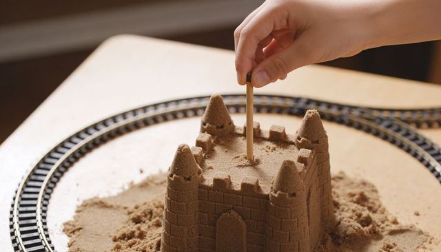 Child placing wooden dowel into detailed indoor sandcastle beside toy train track