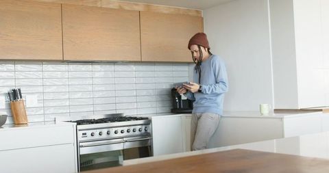 Man wearing beanie standing at modern kitchen counter using tablet near coffee machine