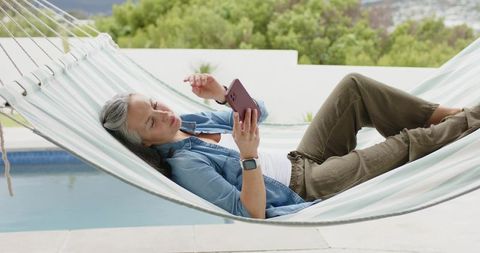 Mature woman relaxing in striped hammock poolside checking smartphone wearing smartwatch