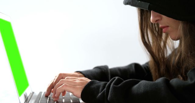 A young Caucasian woman is focused on typing on a laptop with a green screen, with copy space. Her attire suggests a casual or undercover setting, hinting at a narrative of privacy or secrecy.