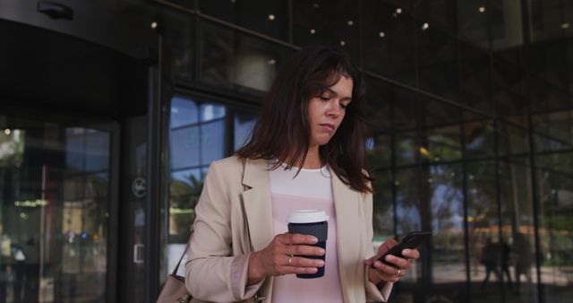 Caucasian businesswoman using smartphone and drinking takeaway coffee outside of modern office. business and business people in office concept.