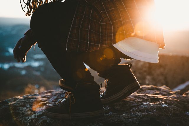 Person wears plaid shirt and boots while squatting on a rocky cliff during sunrise. Sun flare provides a warm glow, creating an adventurous and serene atmosphere. Perfect for content related to outdoor activities, adventure, hiking promotions, lifestyle blogs, and exploring nature.