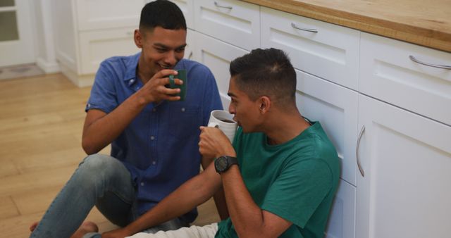 Happy biracial gay male couple sitting on kitchen floor talking and drinking coffee. staying at home in isolation during quarantine lockdown.
